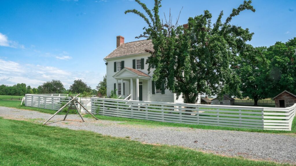 Scenic view of a traditional country house with a white picket fence and lush green surroundings.