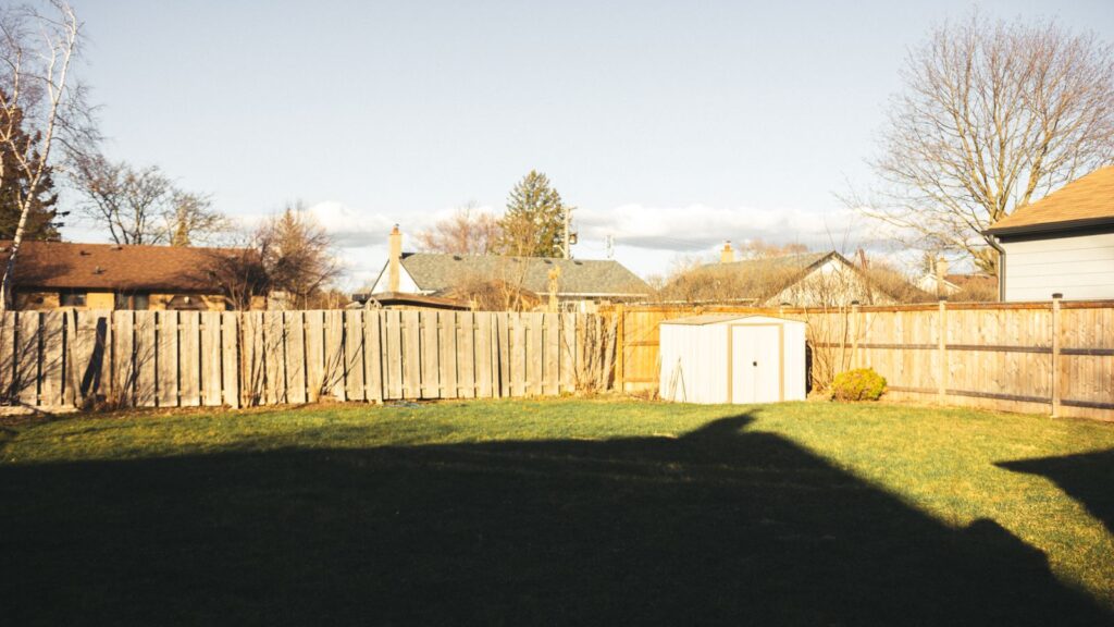 Residential backyard with wooden fence, shed, lawns, and tree shadows on a sunny day.